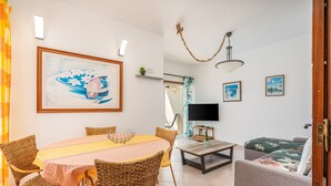 Ein Wohnzimmer mit Esstisch, Korbstühlen, einer Couch, einem Fernseher und Kunstwerken an den Wänden.