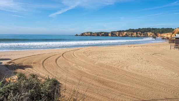 Ein sandiger Strand mit Radspuren, einer Holzkonstruktion und Klippen im Hintergrund.