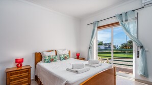 Ein Schlafzimmer mit Bett, Nachttischen, einem Fenster mit Vorhängen und Blick auf eine grüne Landschaft.