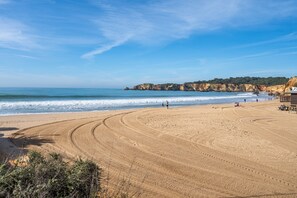 A sandy beach with a curved path leading to the water, a rocky cliff in the background, and a few people walking. - Apartment 'Coral Comfort' with Sea View, Wi-Fi and Air Conditioning (Portimão)