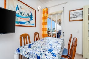 A dining area with a table set for four, wooden chairs, a TV mounted on the wall, and a view through a glass door to the outside.