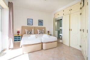 Une chambre à coucher avec un lit double, deux tables de chevet, une armoire et une salle de bain visible à travers une porte ouverte.