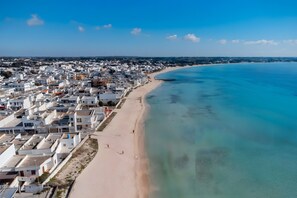 Vue aérienne d’une ville côtière avec une plage, des bâtiments blancs et une eau d’un bleu limpide.