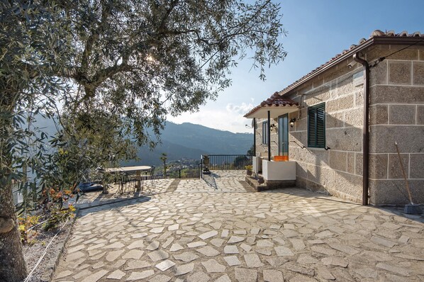 A stone patio with a table and chairs, a small building with shutters, and a view of mountains in the background.