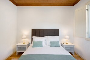 Quarto com uma cama, duas mesas de cabeceira com luminárias, teto de madeira e paredes brancas.