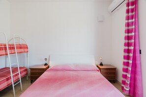 Een slaapkamer met een stapelbed, een eenpersoonsbed, een nachtkastje met een lamp en een luchtverkoeler aan de muur.