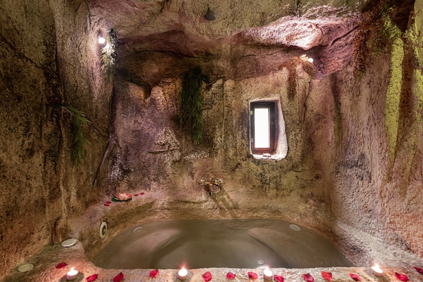 Una habitación con forma de cueva, una bañera, un ventanal pequeño y velas.
