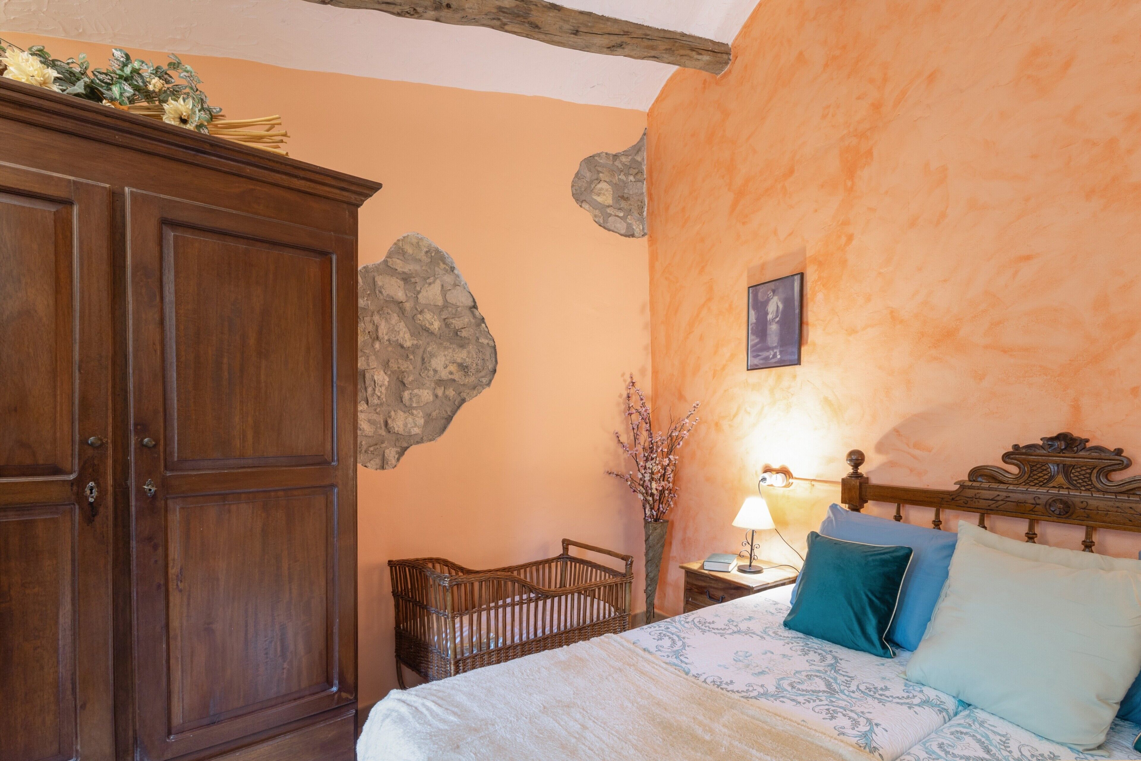 A bedroom with a wooden wardrobe, a bed with patterned bedding, a nightstand with a lamp, and a wall-mounted picture.