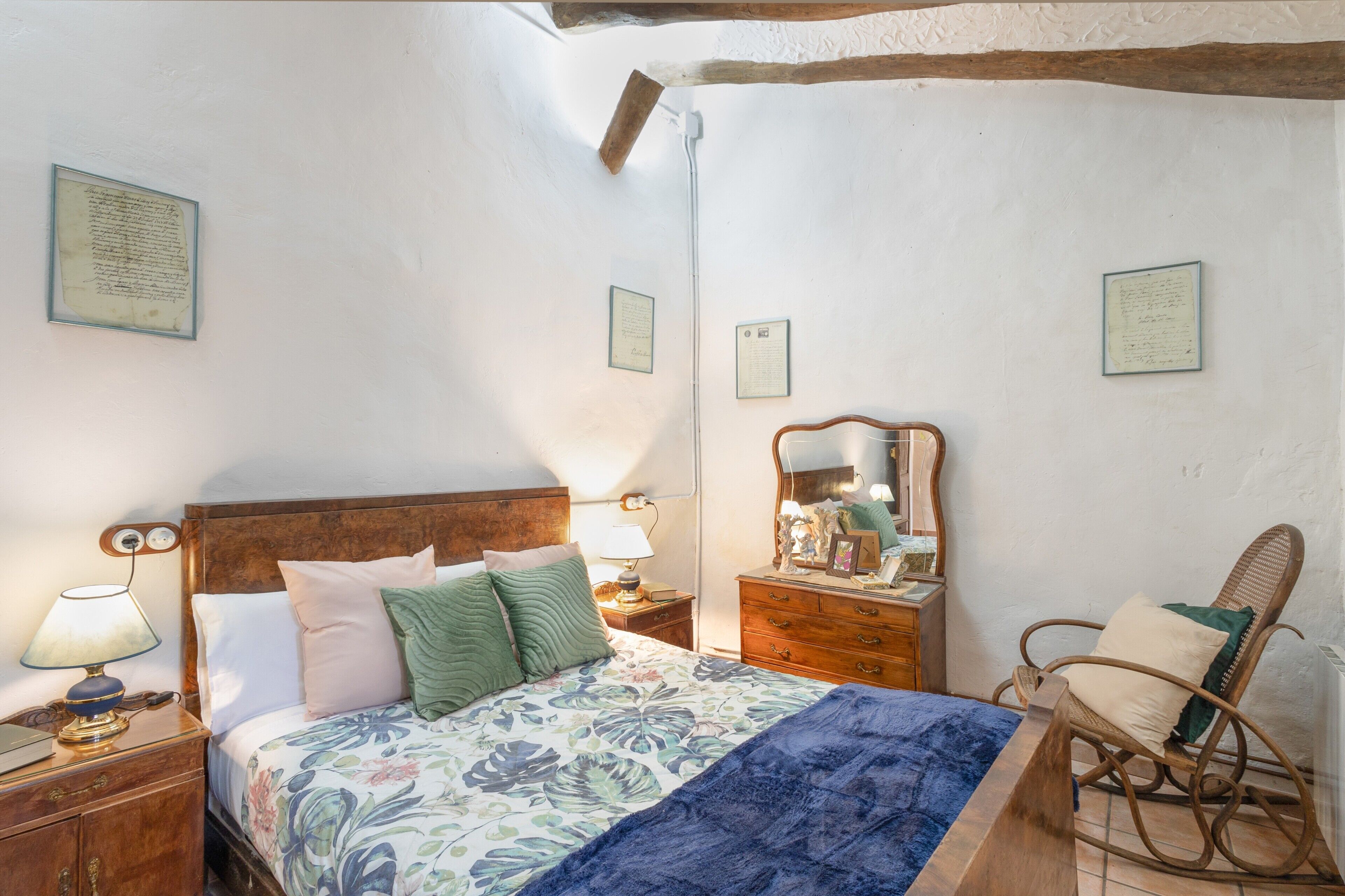 Un dormitorio con una cama de madera, una mesita de noche con lámpara, un aparador, un sillón mecedora y documentos enmarcados en la pared.