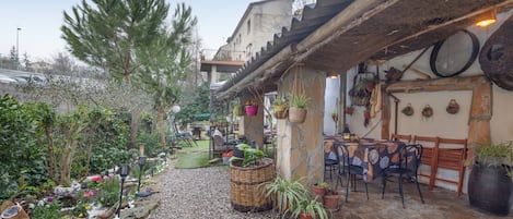Un patio con suelo de piedra, plantas en macetas, mobiliario exterior y un edificio rústico de techo inclinado.