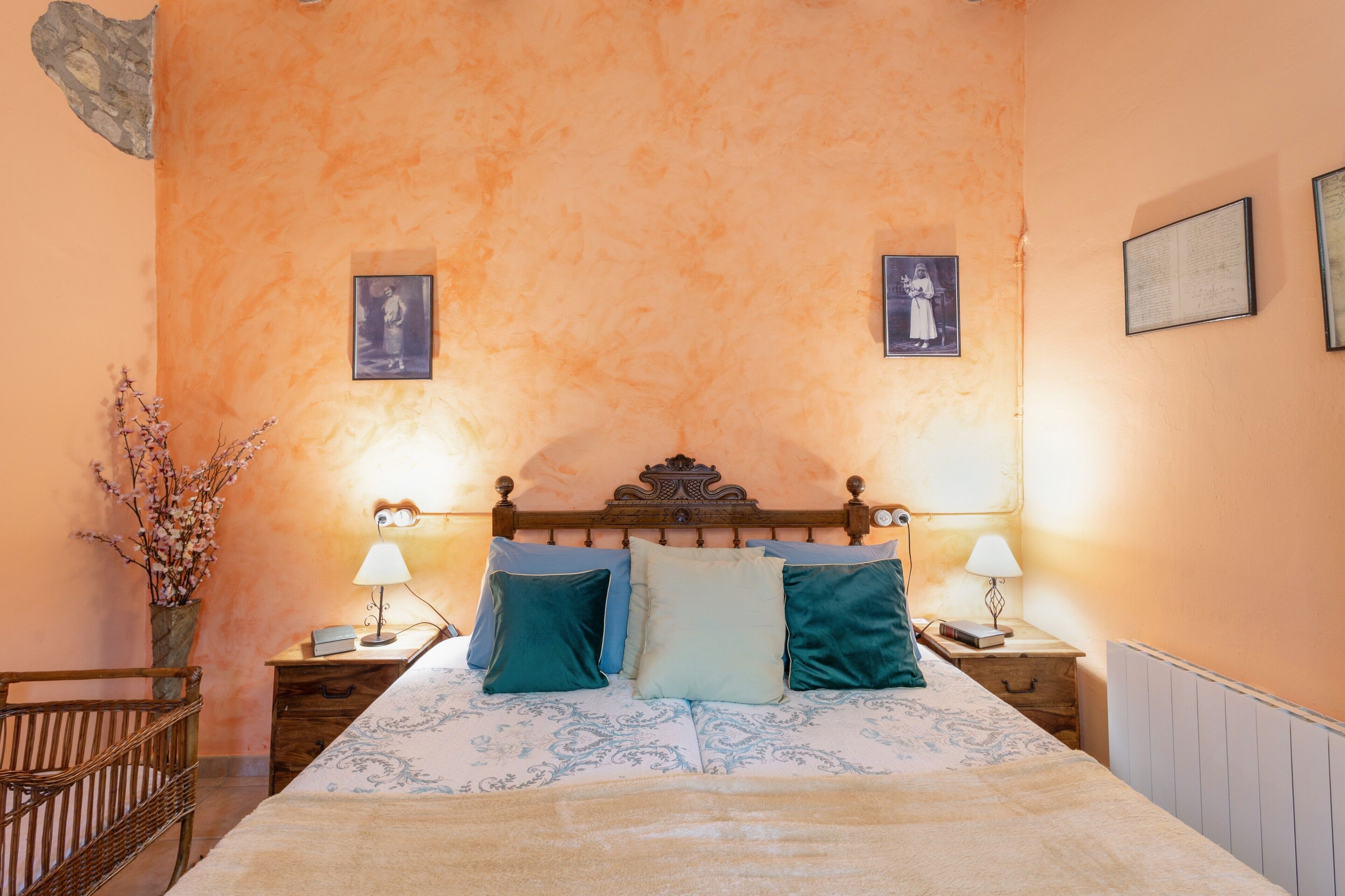 Een slaapkamer met een houten hoofdbord, een bed met beddengoed met een patroon, twee nachtkastjes met lampen, en een vaas met takken aan de linkerkant.