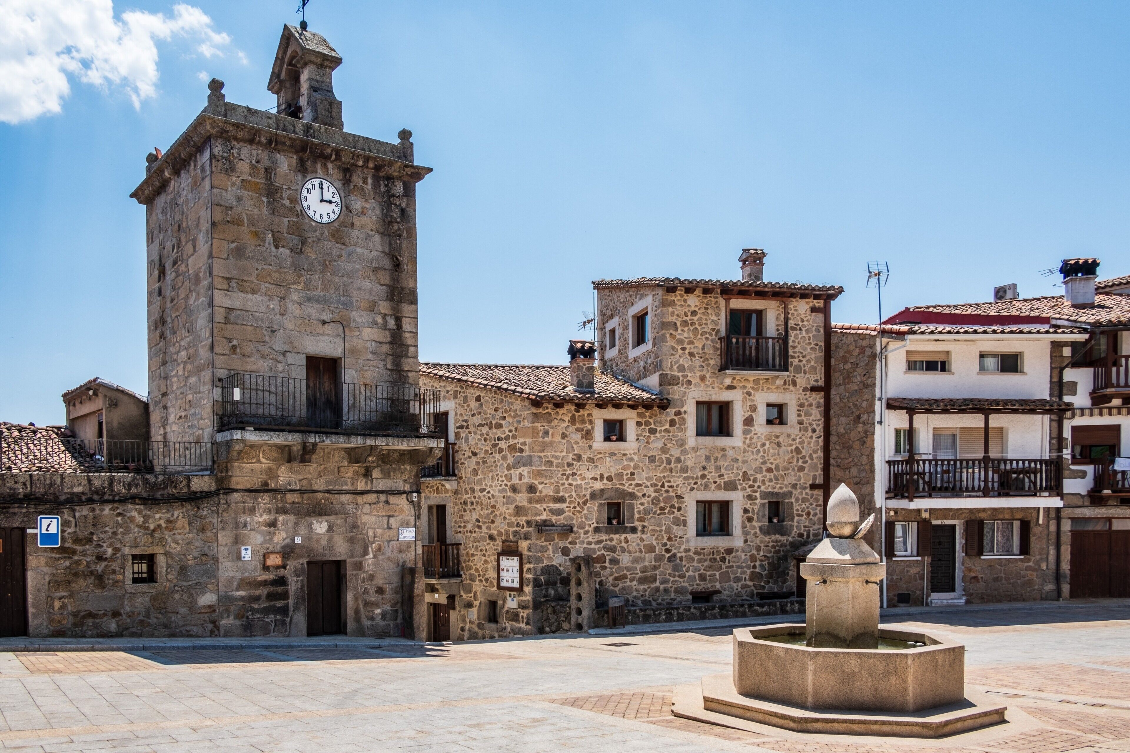 A historic stone building with a clock tower, a fountain, and a clear blue sky.