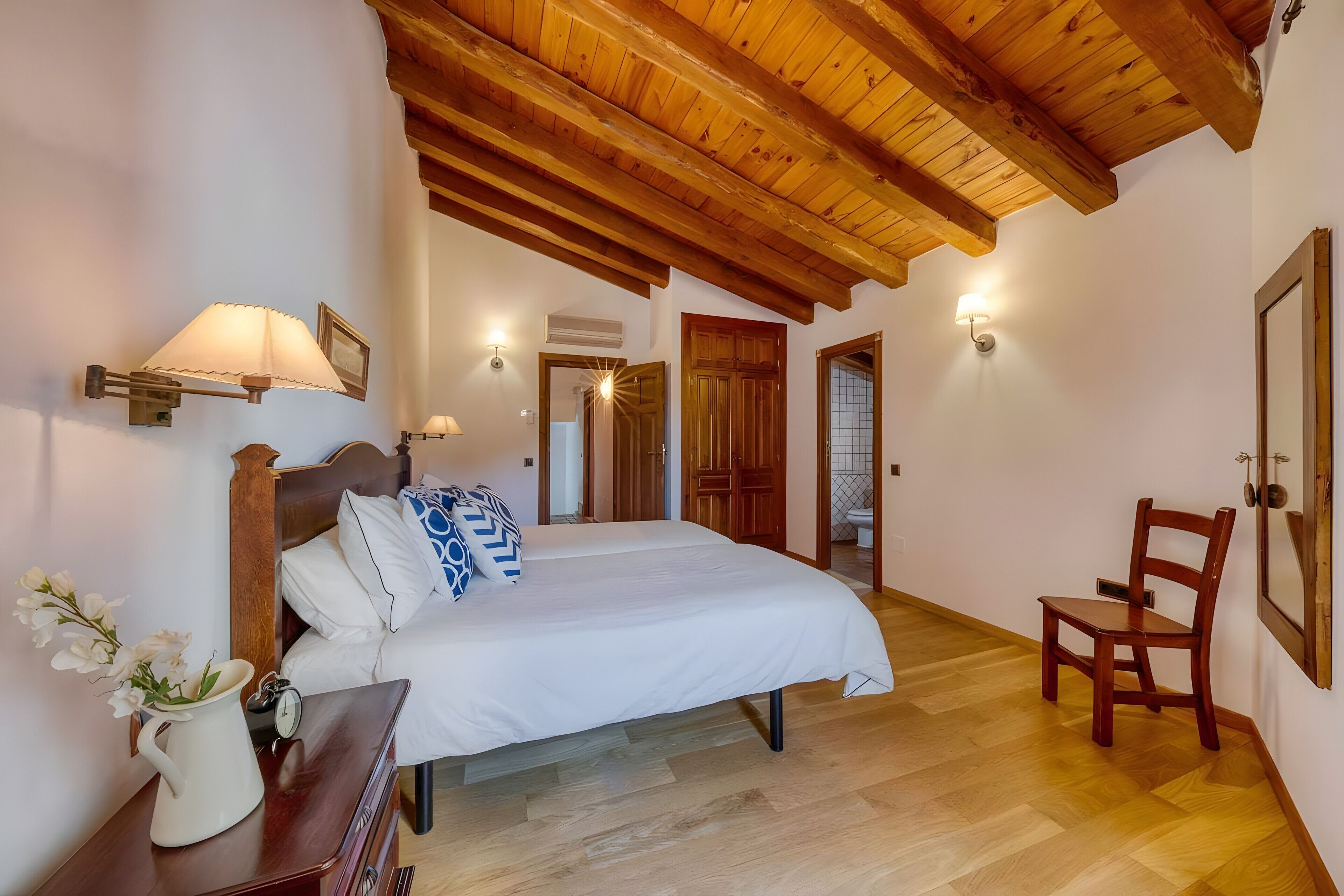 A bedroom with a wooden ceiling, a bed with white bedding, a wooden chair, and wall-mounted lights.