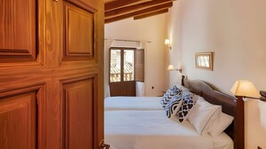 Habitación de hotel con puerta de madera, cama con almohadas estampadas, mesita de noche con lámpara y vistas al exterior.