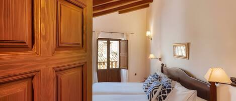 A hotel room with a wooden door, a bed with patterned pillows, a nightstand with a lamp, and a view of the outside.