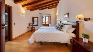 Un dormitorio con vigas de madera, una cama con ropa de cama blanca, una mesita de noche, una silla y una ventana con cortinas.