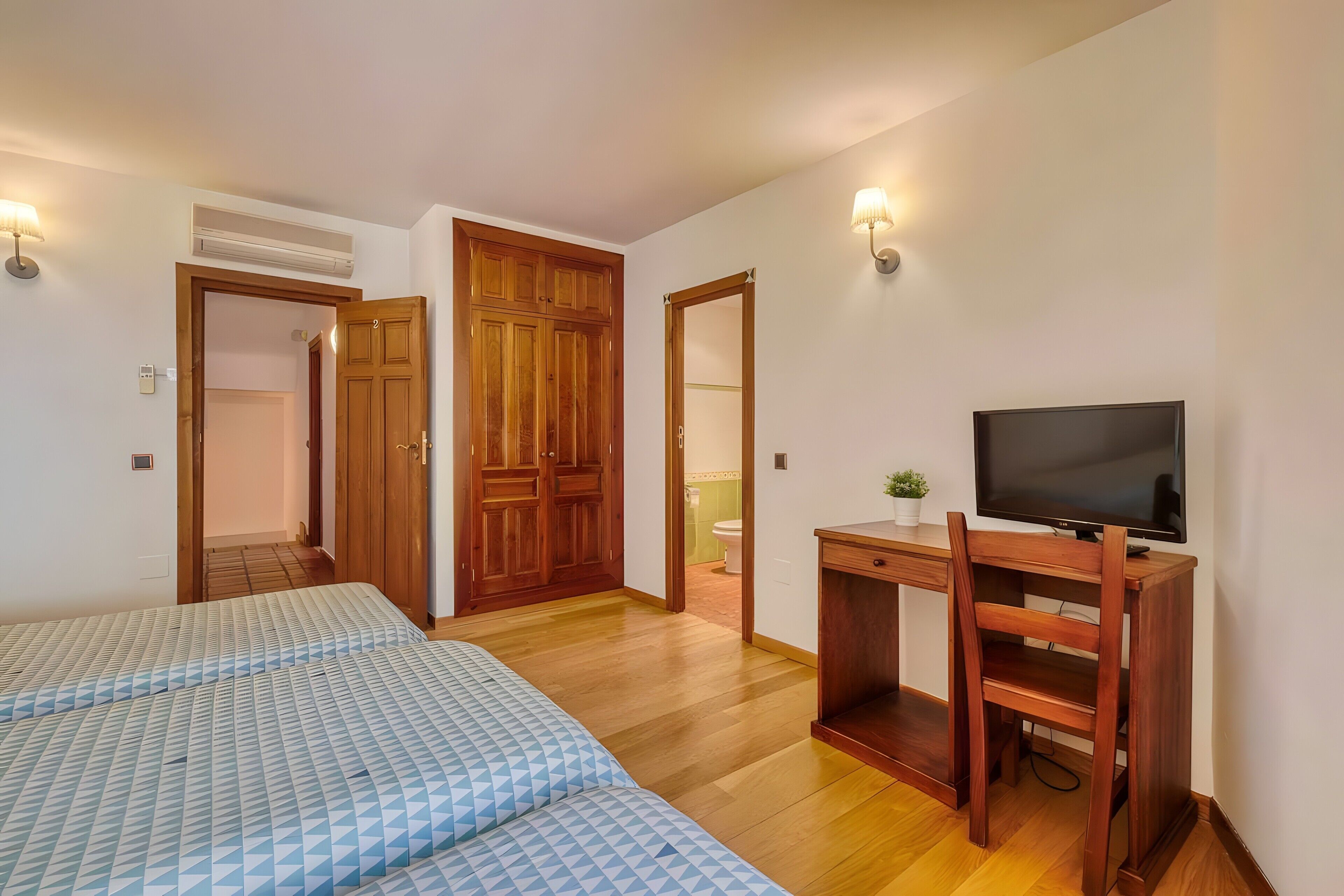 Un dormitorio con dos camas, un escritorio de madera y un televisor, y un baño visible a través de una puerta abierta.