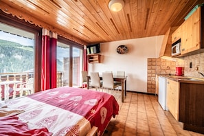 A room with a bed, a dining table with chairs, a kitchen area, and a view of the outdoors.