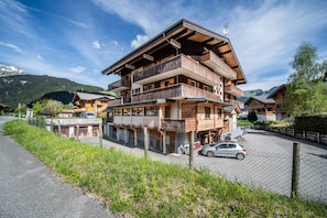 A multi-story wooden building with balconies, a parking area, and a mountain backdrop.
