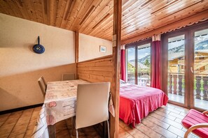 A wooden interior with a dining table, chairs, a bed with a red bedspread, and a view of mountains through large windows.