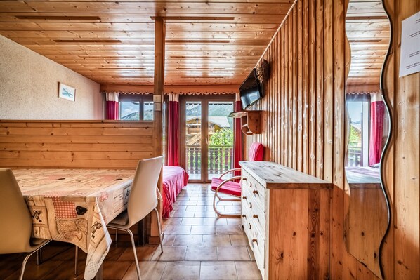 A wooden interior with a dining table, chairs, a TV mounted on the wall, and a view through a glass door.