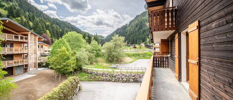 Un edificio de madera con balcón, un muro de piedra y vistas de un valle con árboles y una montaña.