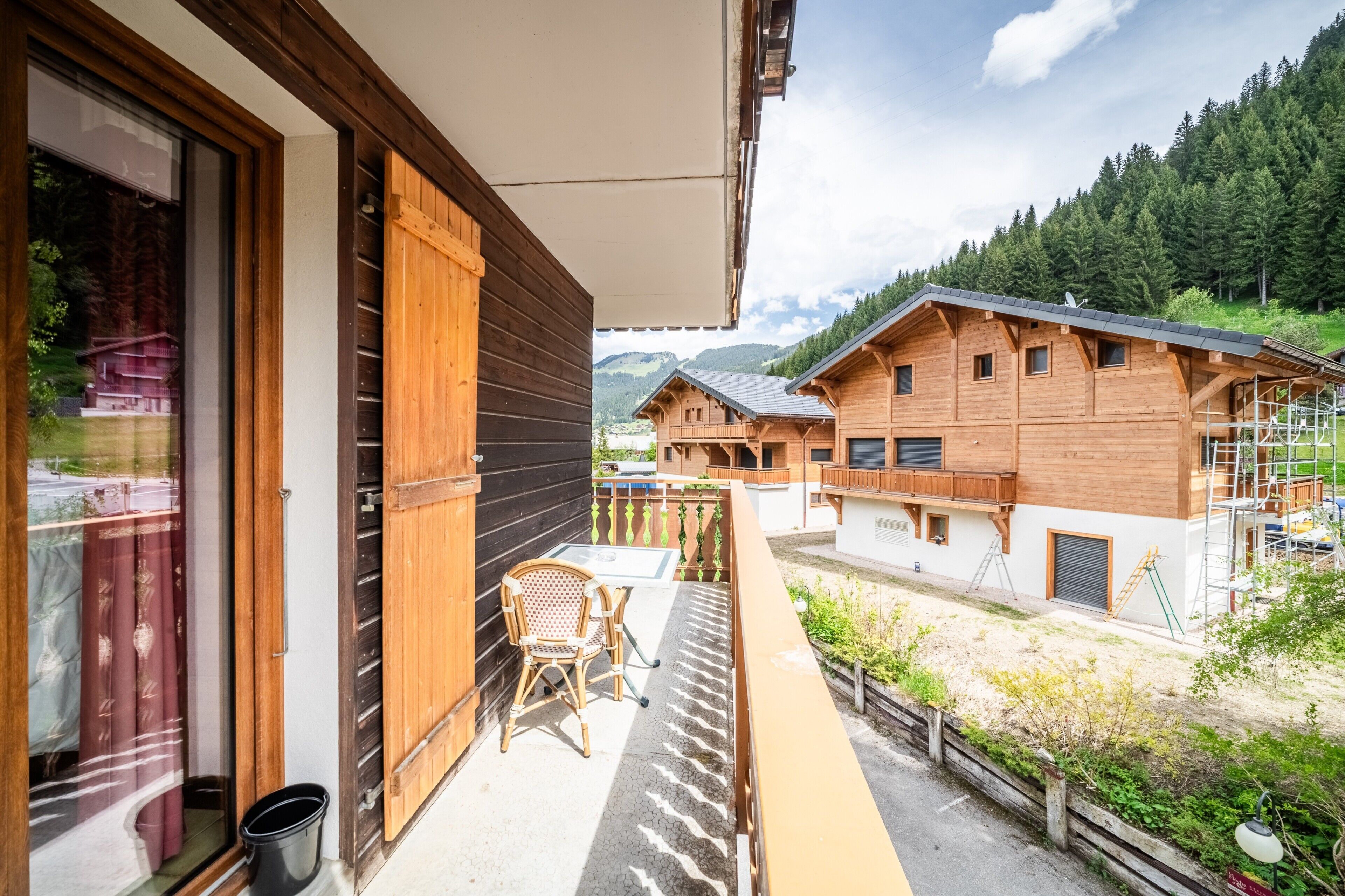 A wooden balcony with a table and chair, overlooking a residential area with houses and mountains.