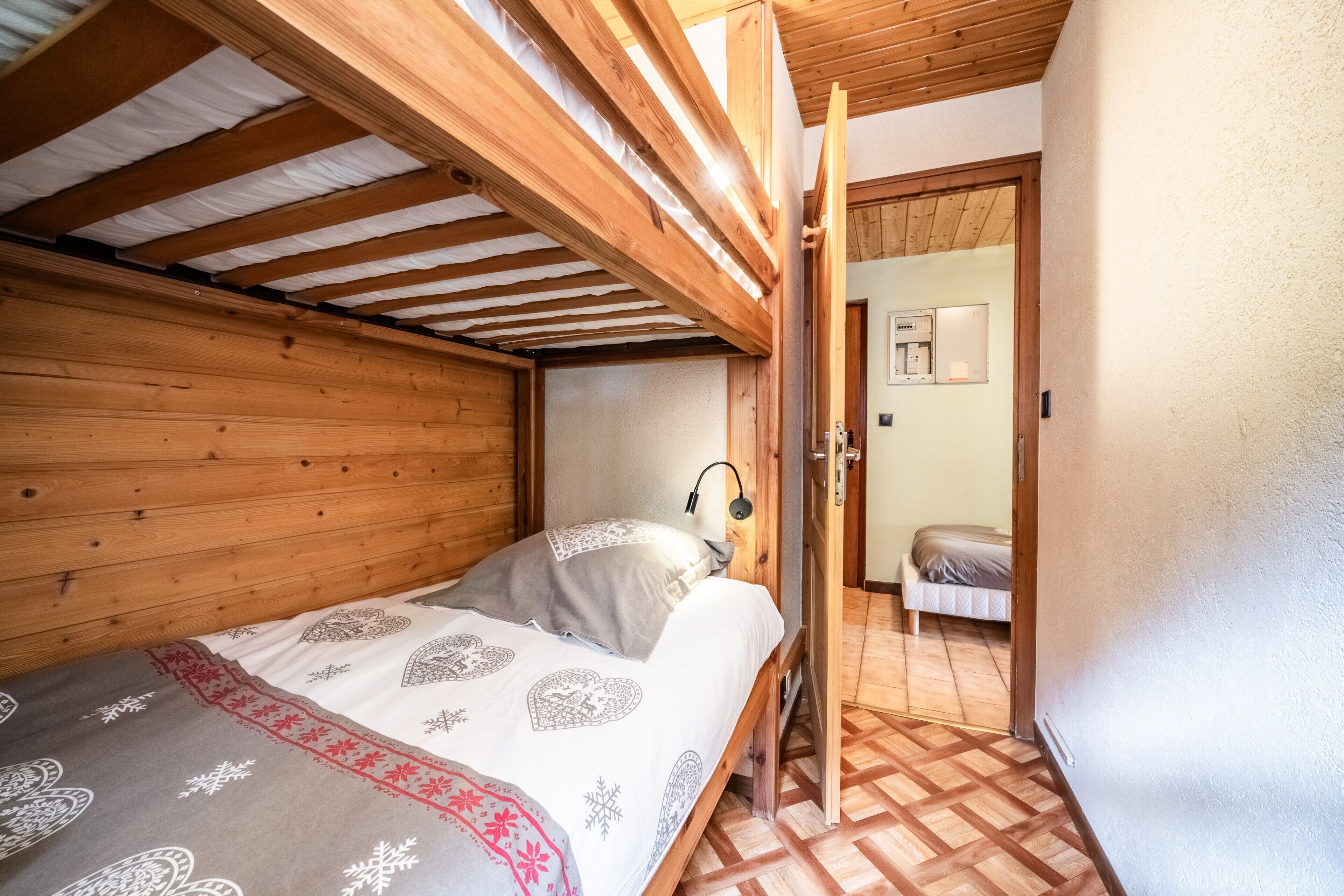 A bedroom with a wooden ceiling, a bed with patterned bedding, a bedside lamp, and a view into an adjacent room.