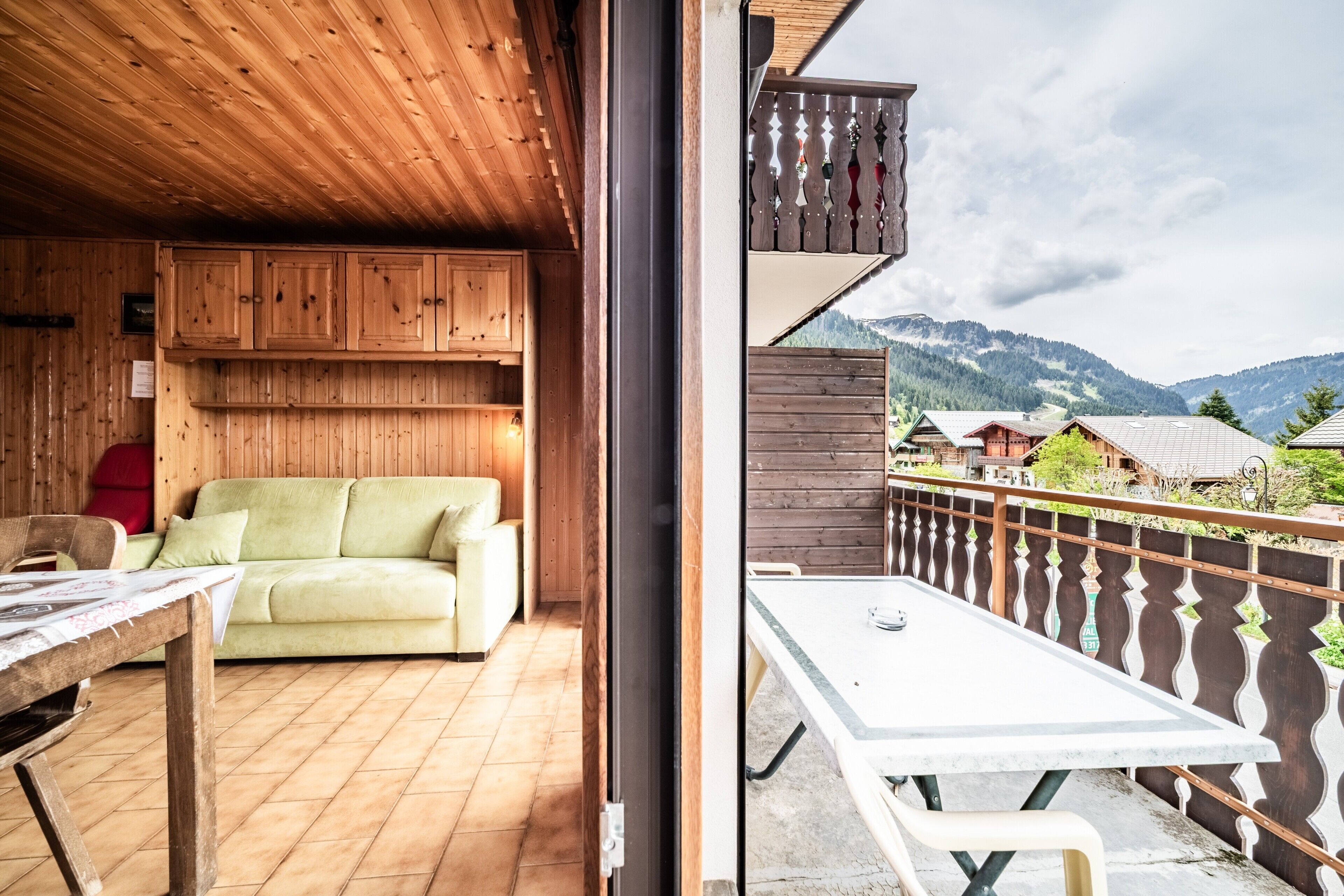 A wooden interior with a sofa and a table, a balcony with a railing, and a mountainous landscape in the background.