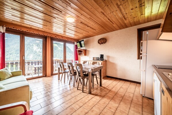 A room with a wooden ceiling, a dining table with chairs, a sofa, a kitchen area with a refrigerator, and a TV mounted on the wall.