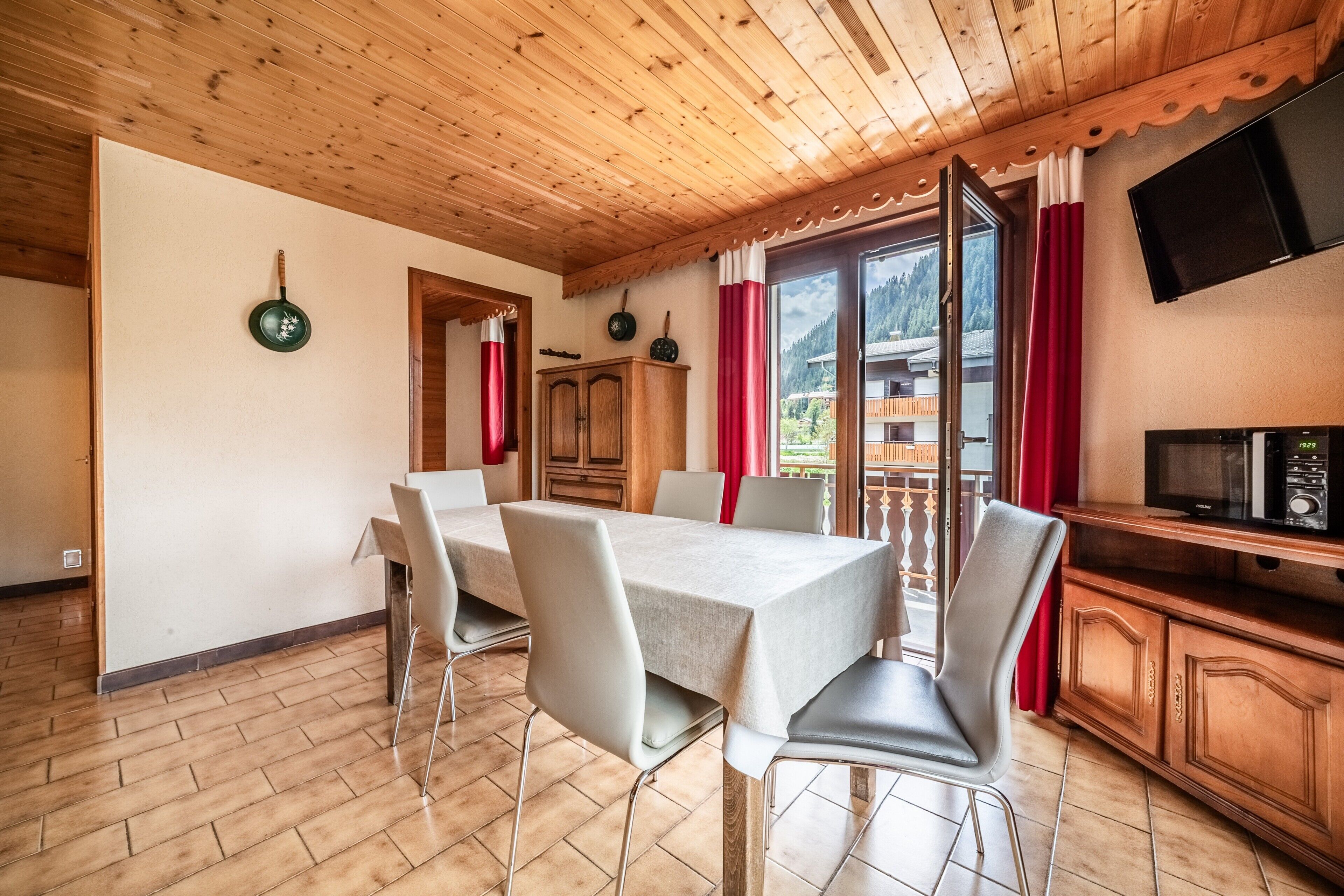 Un comedor con mesa y sillas, armarios de madera, un televisor y vista al exterior.