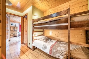 A bunk bed with a wooden headboard, a bedside table with a lamp, and a view into a room with a wooden floor and a cabinet.