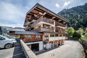 Un edificio de madera de varios pisos con balcones, un estacionamiento y montañas al fondo.