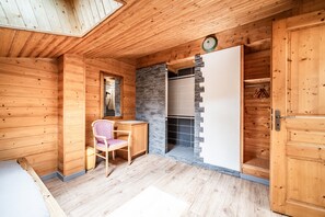 A wooden cabin interior with a bed, a chair, a desk, a stone fireplace, and a wardrobe.