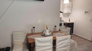 A dining area with a table set for two, covered chairs, a TV mounted on the wall, and a kitchen in the background. - Studio Apartment 'Corfu Oasis' with Mountain View, Wi-Fi and Air Conditioning (Corfu)
