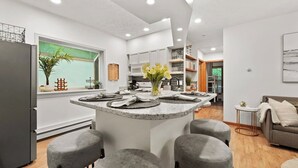 Eine moderne Küche mit einer zentralen Kochinsel, grauen Barhocker und Blick auf den Wohnbereich mit einem grauen Sofa und einem kleinen runden Tisch.
