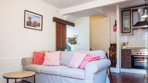 Un salon avec un canapé, une petite table ronde, un coin cuisine avec des placards et un four, et un tableau encadré au mur.
