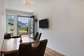 Un coin repas avec une table et des chaises en bois, un téléviseur fixé au mur et une vue sur les montagnes par la porte vitrée.