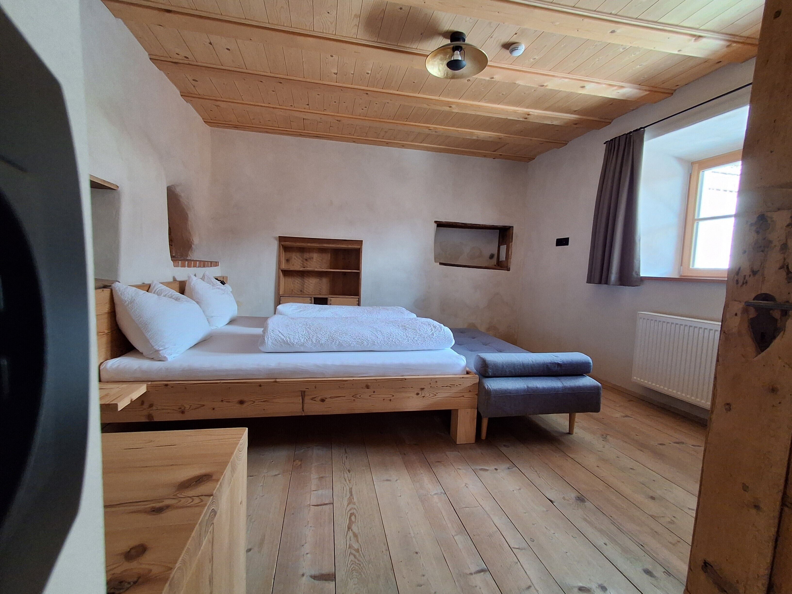 Ein Zimmer mit Holzboden, einem Bett, einer kleinen Ablage, einem Fenster mit Vorhängen und einem Heizkörper.