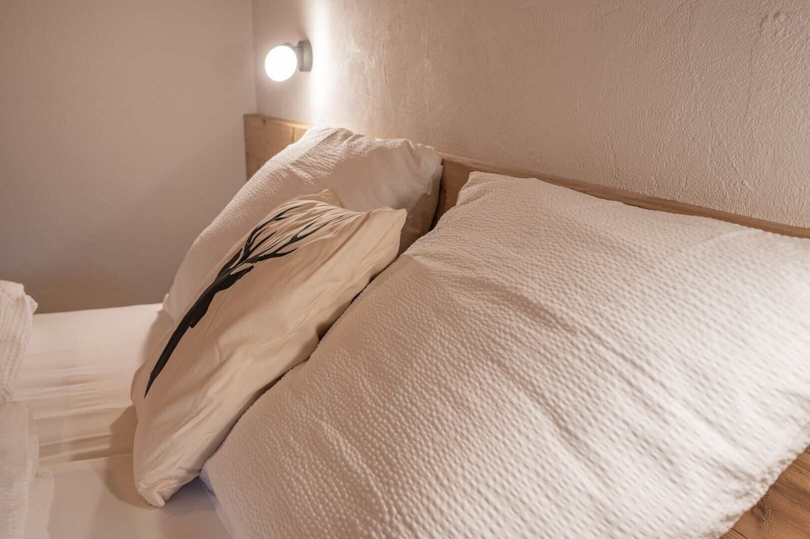 Un lit bien fait, avec du linge de lit blanc et un seul coussin décoratif orné d’un motif représentant un arbre noir.