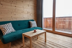 Una habitación con un sofá color turquesa, una mesa de madera y un ventanal con vistas a una terraza de madera.