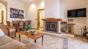 A living room with a fireplace, a television mounted on the wall, wicker furniture, and a dining area. - Holiday Home 'León' with Private Terrace, Wi-Fi and Air Conditioning (Almodóvar del Río)