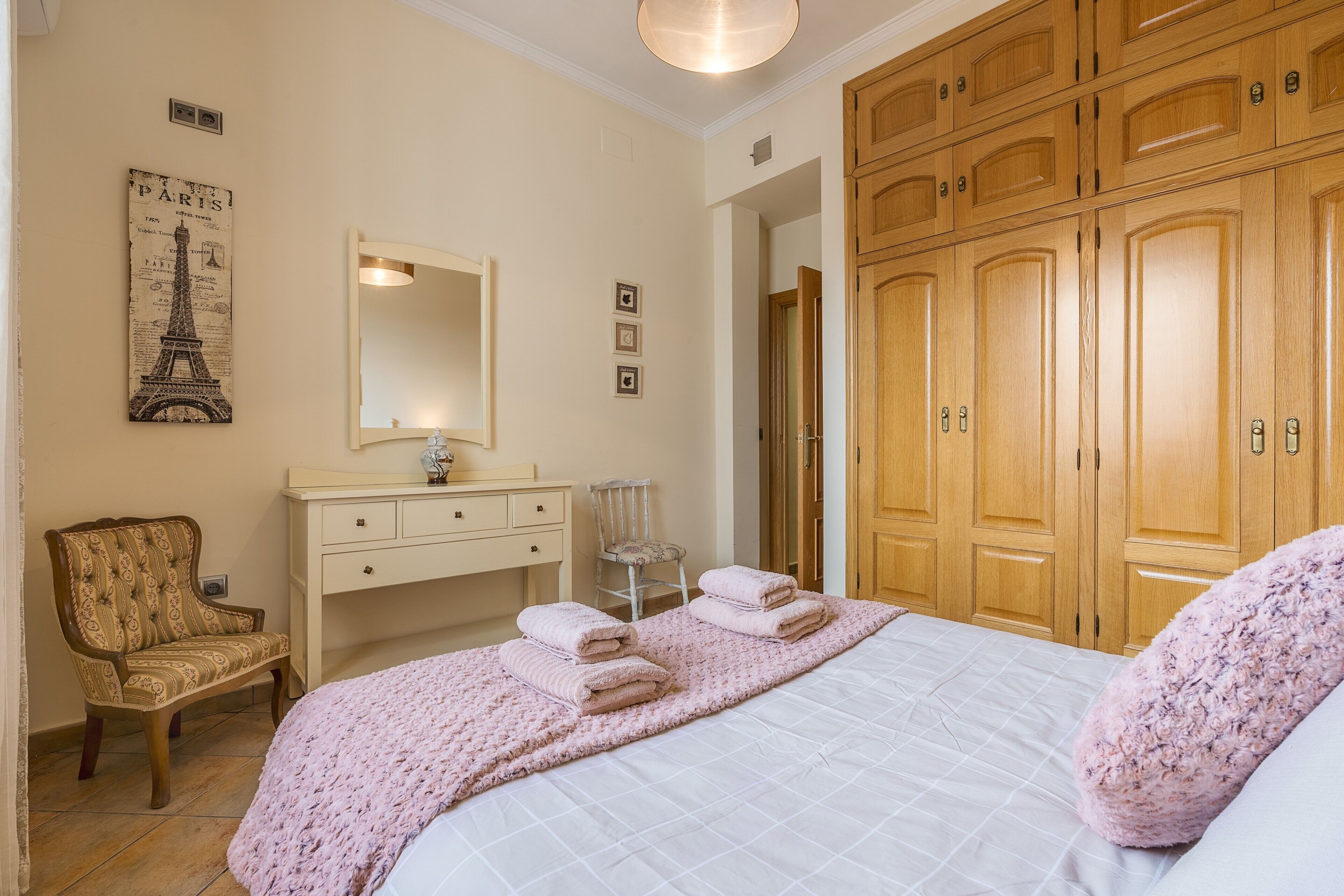 A bedroom with a bed, a wooden wardrobe, a chair, a dresser, and a wall-mounted mirror.