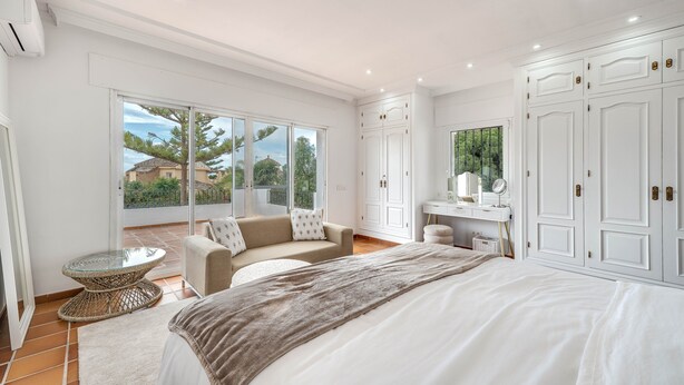 A bedroom with a large bed, a sofa, a small table, a wardrobe, and a bathroom with a mirror.