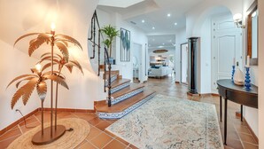 A spacious hallway with a palm tree floor lamp, a staircase with patterned steps, and a large rug.