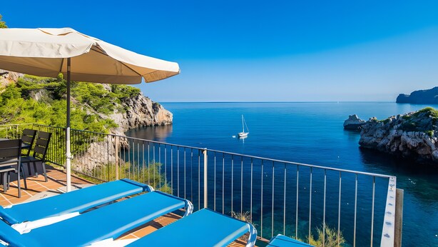A balcony with blue lounge chairs and a beige umbrella overlooking a calm sea with a sailboat. - Holiday Home 'Cala Deia' with Sea View, Wi-Fi and Air Conditioning (Illes Balears)