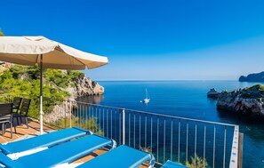 A balcony with blue lounge chairs and a beige umbrella overlooking a calm sea with a sailboat. - Holiday Home 'Cala Deia' with Sea View, Wi-Fi and Air Conditioning (Illes Balears)