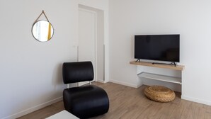 Un salon moderne avec un fauteuil en cuir noir, un téléviseur à écran plat posé sur une étagère en bois, un miroir rond accroché au mur et un panier en osier sur le sol.