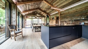 A spacious kitchen with a large island, wooden ceiling beams, and a view through large windows.