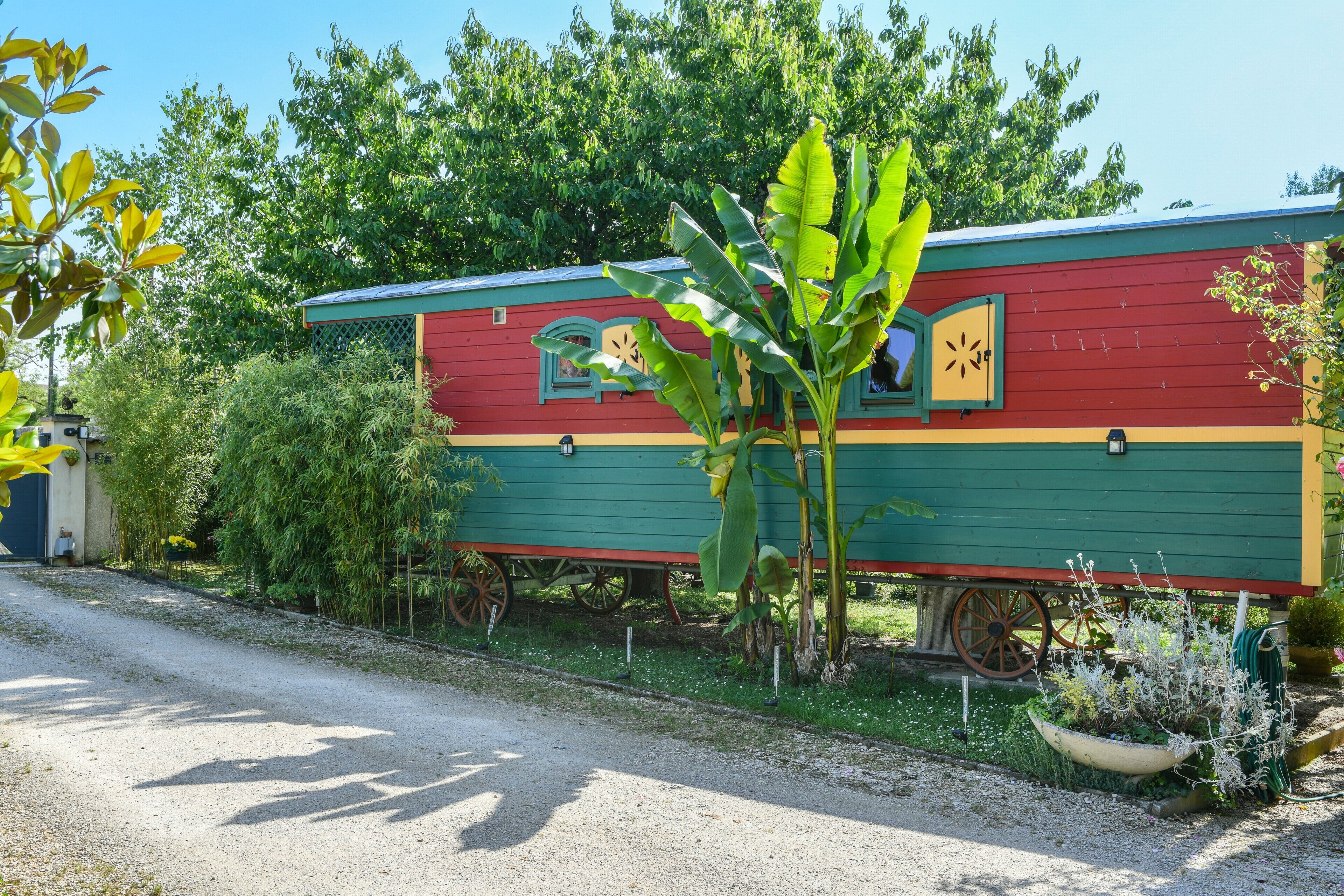 A stationary, colorful RV with a green and red exterior, positioned on a gravel path with trees and a planter nearby.